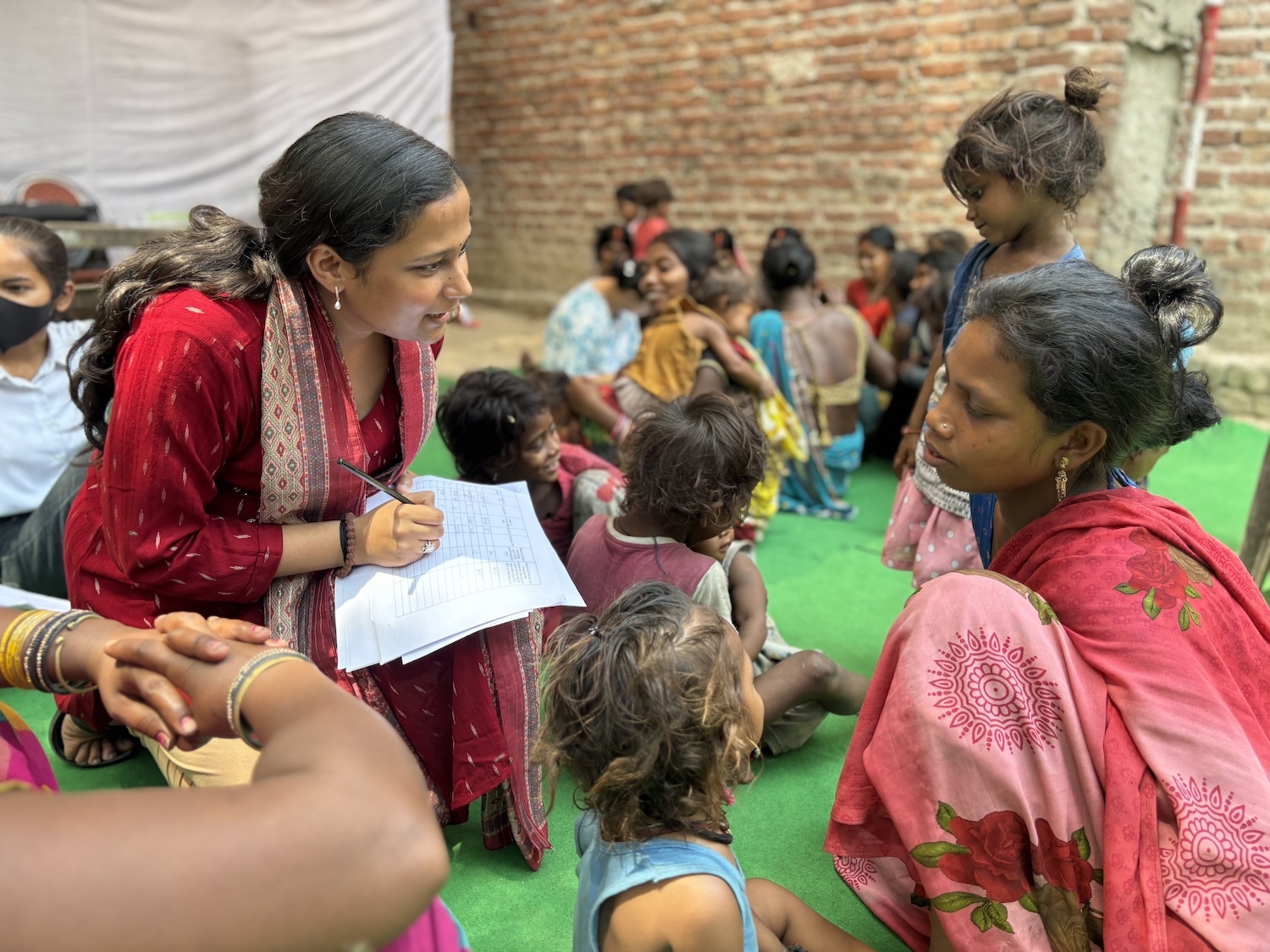 A woman in traditional attire is discussing the Period Awareness Drive with a seated woman and children outdoors. She is holding a paper and pen, seemingly taking notes, amidst a group of people, including children, in the background. This initiative is supported by GPF India to promote sustainable solutions.