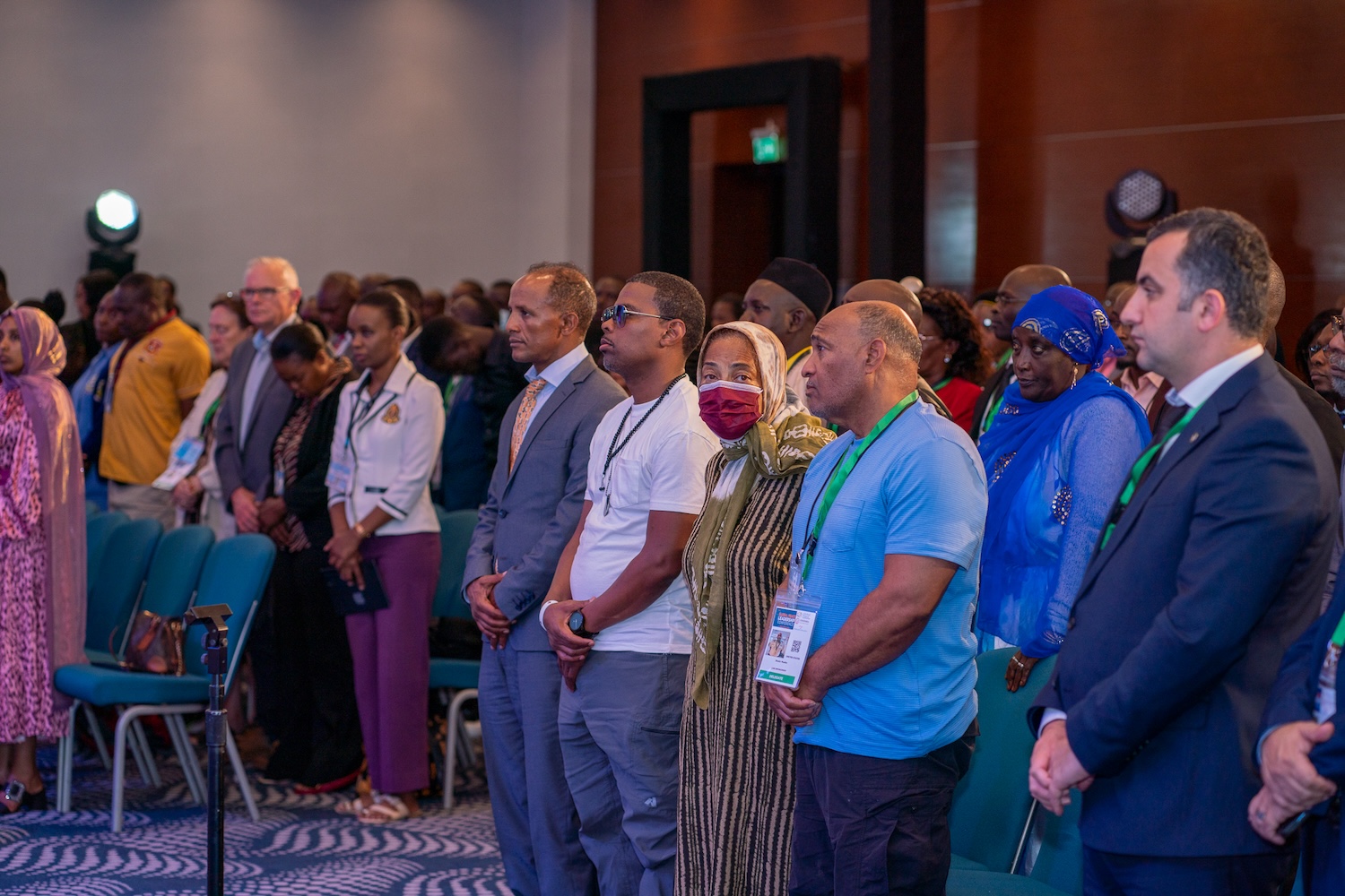 A diverse group of people, standing in rows, attends a formal event in a conference room during GPLC Africa in Kenya. Some individuals are dressed in traditional attire while others wear business or casual clothing, united by a shared moment of prayer.