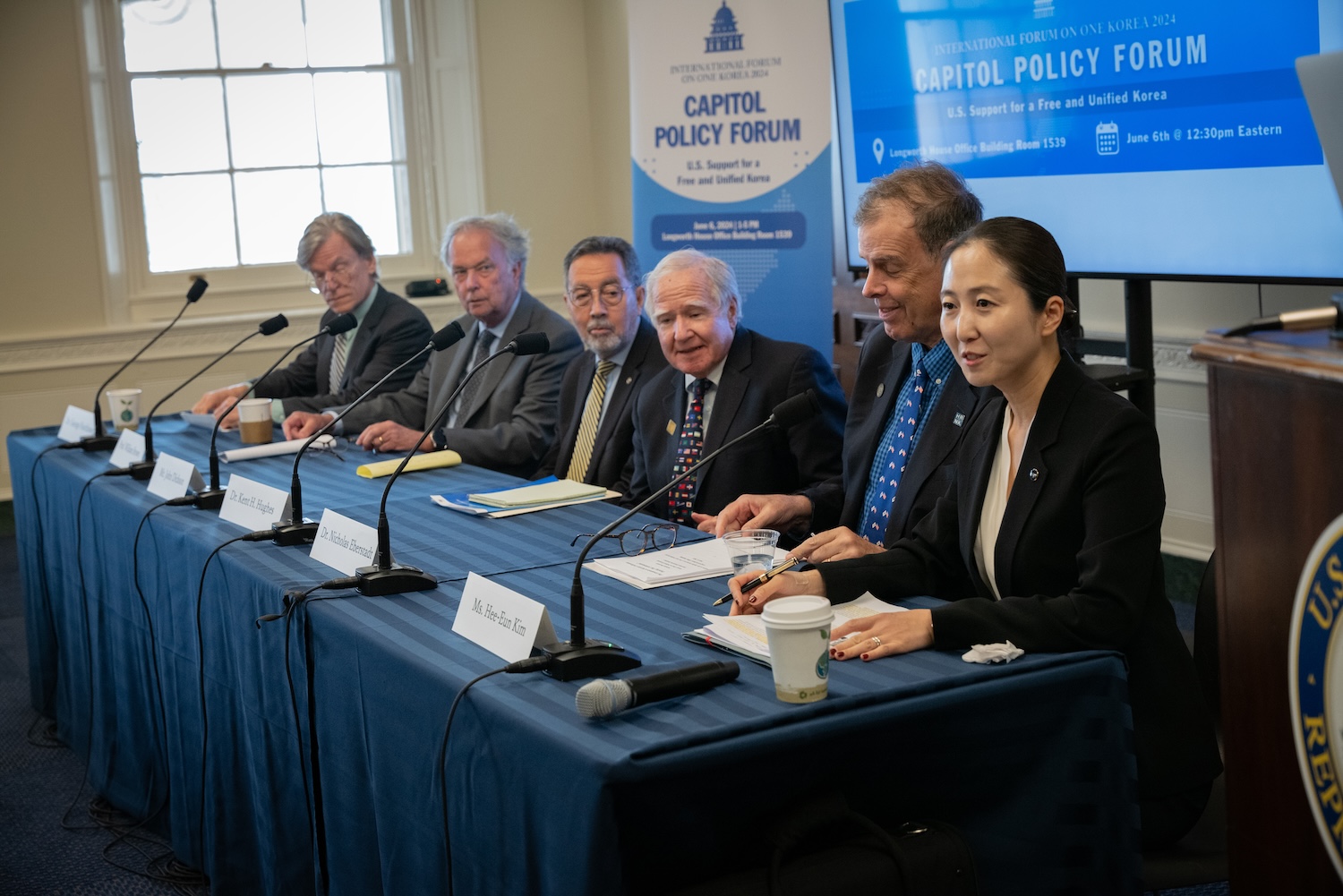 Six individuals sit at a panel table with microphones and nameplates in front of them, participating in the Capitol Policy Forum. A blue Capitol Policy Forum banner is displayed in the background, announcing an International Forum on policy issues impacting various regions, including a Unified Korea.