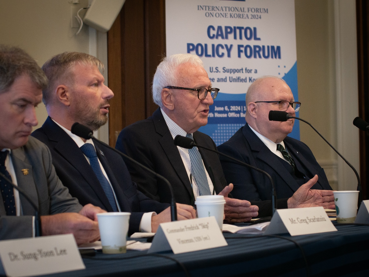 Four men in suits are seated at a table with microphones, papers, and coffee cups. Behind them is a banner for the "Capitol Policy Forum" focusing on U.S. support for a Unified Korea.