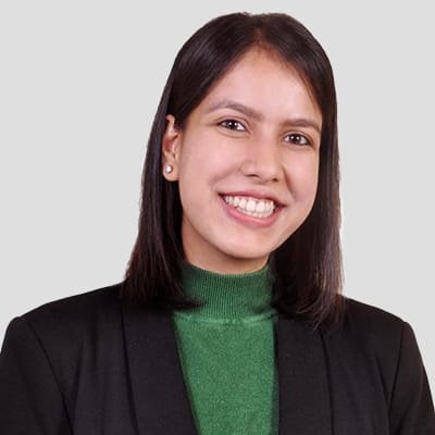 Stella, smiling brightly in a green turtleneck and black blazer, stands against a plain background, exuding confidence and style.