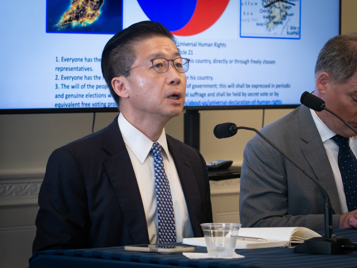 A man in a dark suit and polka dot tie speaks at an International Forum, seated next to another man in a gray suit. A presentation slide with text and images about One Korea is displayed in the background.