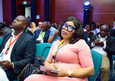 A group of people are seated in an indoor setting, some taking notes and others using their phones. One woman in the foreground, possibly a participant in the Environmental Track of GPLC Africa 2024, is wearing large glasses and holding a notebook.