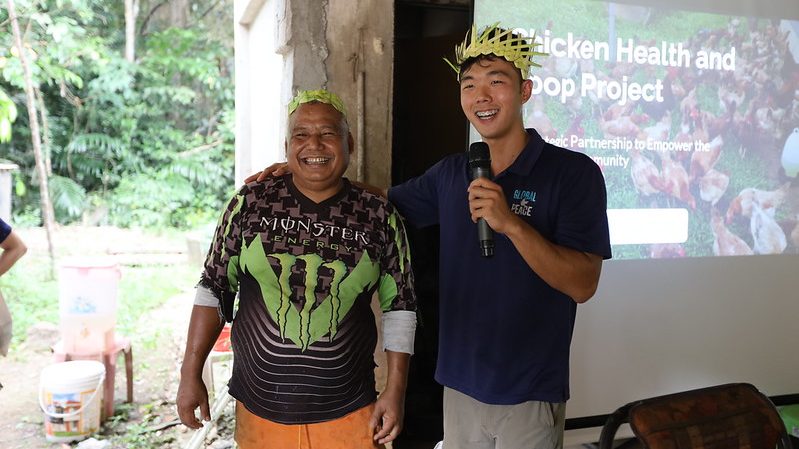 Two men standing outdoors; one is smiling and wearing a Monster Energy shirt, the other is speaking into a microphone. Both are wearing leaf hats. A screen featuring text about GPF Malaysia's initiatives in rural communities is visible in the background.