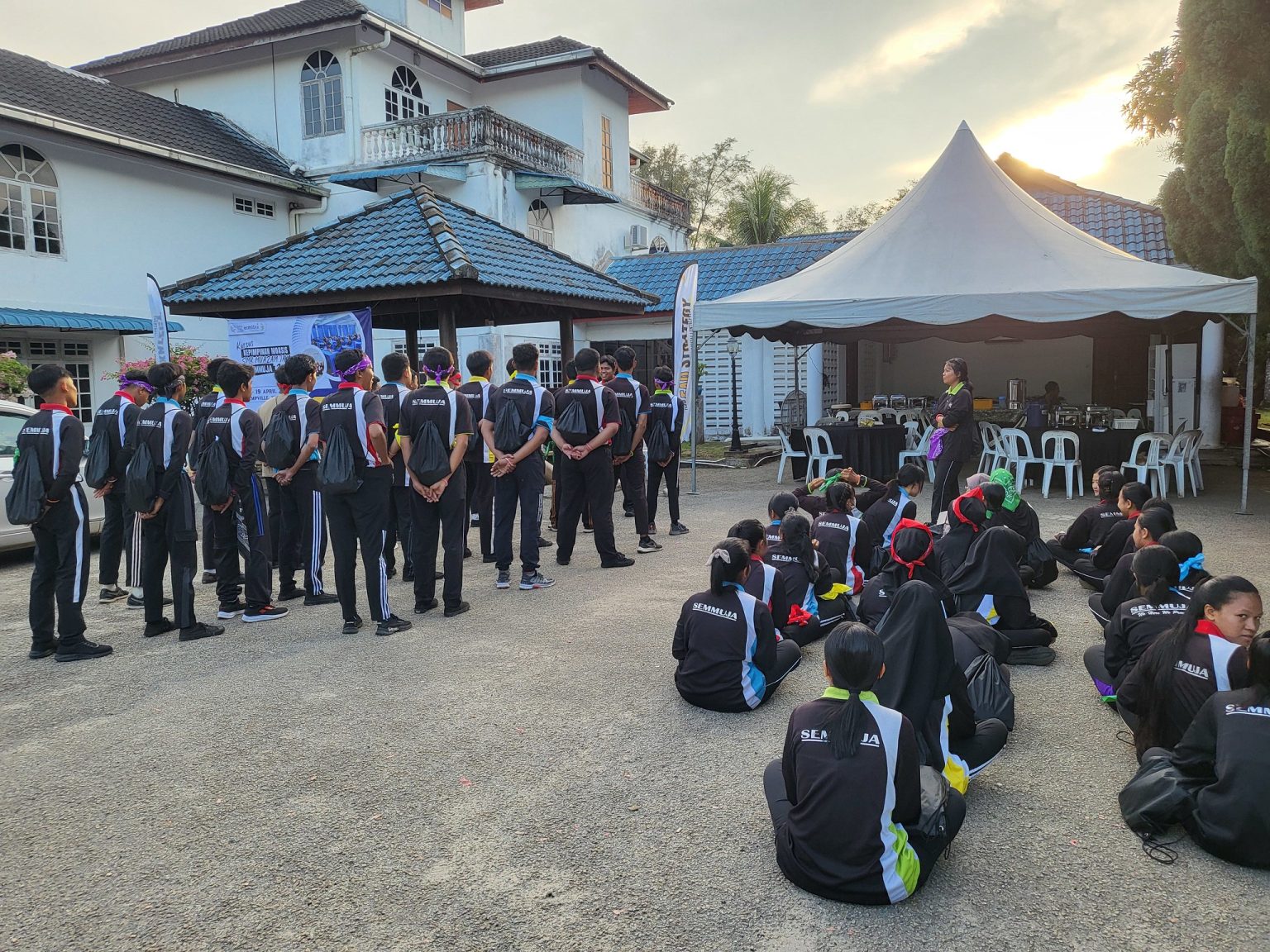 A group of people in matching uniforms are gathered outdoors near a building, fostering an empowering atmosphere. Some are standing in lines while others are seated. There is a tent and several empty chairs nearby, embodying the unity often seen at events focused on Indigenous Youth in Malaysia.