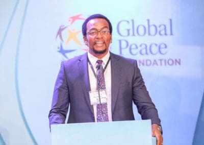 A man in a suit and tie stands at a podium speaking, with a "Global Peace Foundation" logo in the background, addressing attendees of the GPLC Africa 2024 Education Track.