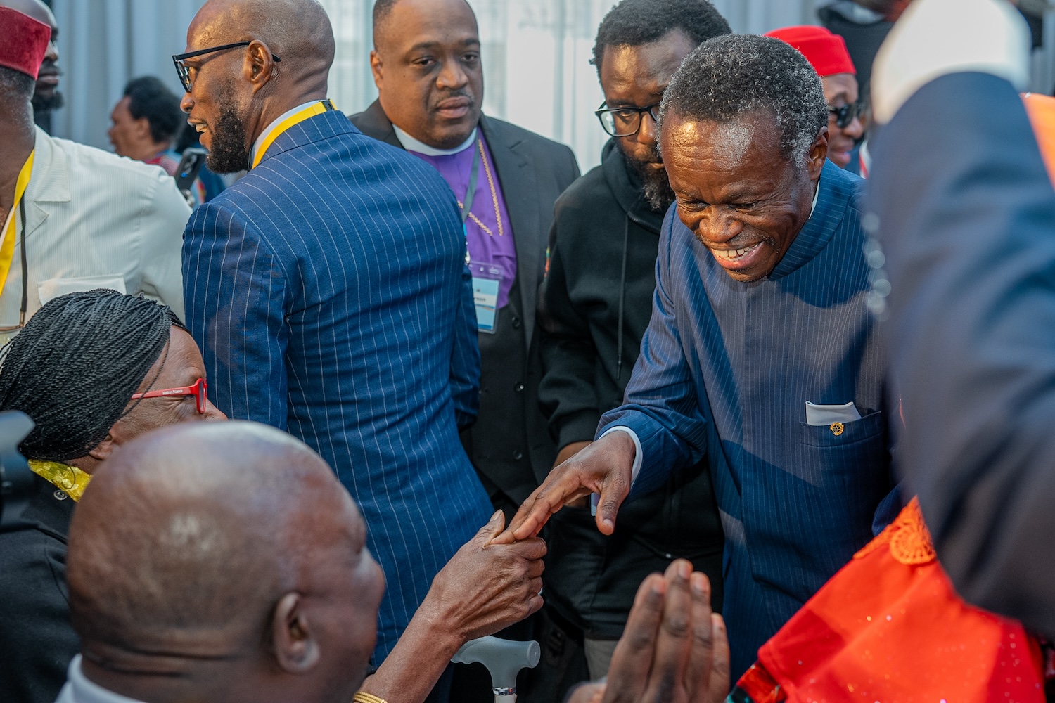A group of well-dressed individuals interacts at an event. One man smiles and shakes another person's hand while others converse in the background, creating a warm atmosphere reminiscent of the Ubuntu Track at GPLC Africa 2024.