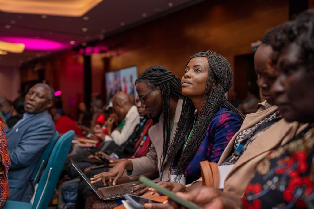 A group of people sit attentively in a conference room at the GPLC Africa 2024 plenary, with some taking notes on laptops and others looking forward. The setting appears formal and focused.