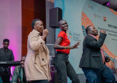 Four people on stage are singing and dancing in front of a backdrop that reads "One Family Under God." A keyboard player is visible in the background. The event appears to be organized by the Global Peace Foundation as part of the Africa 2024 Plenary.