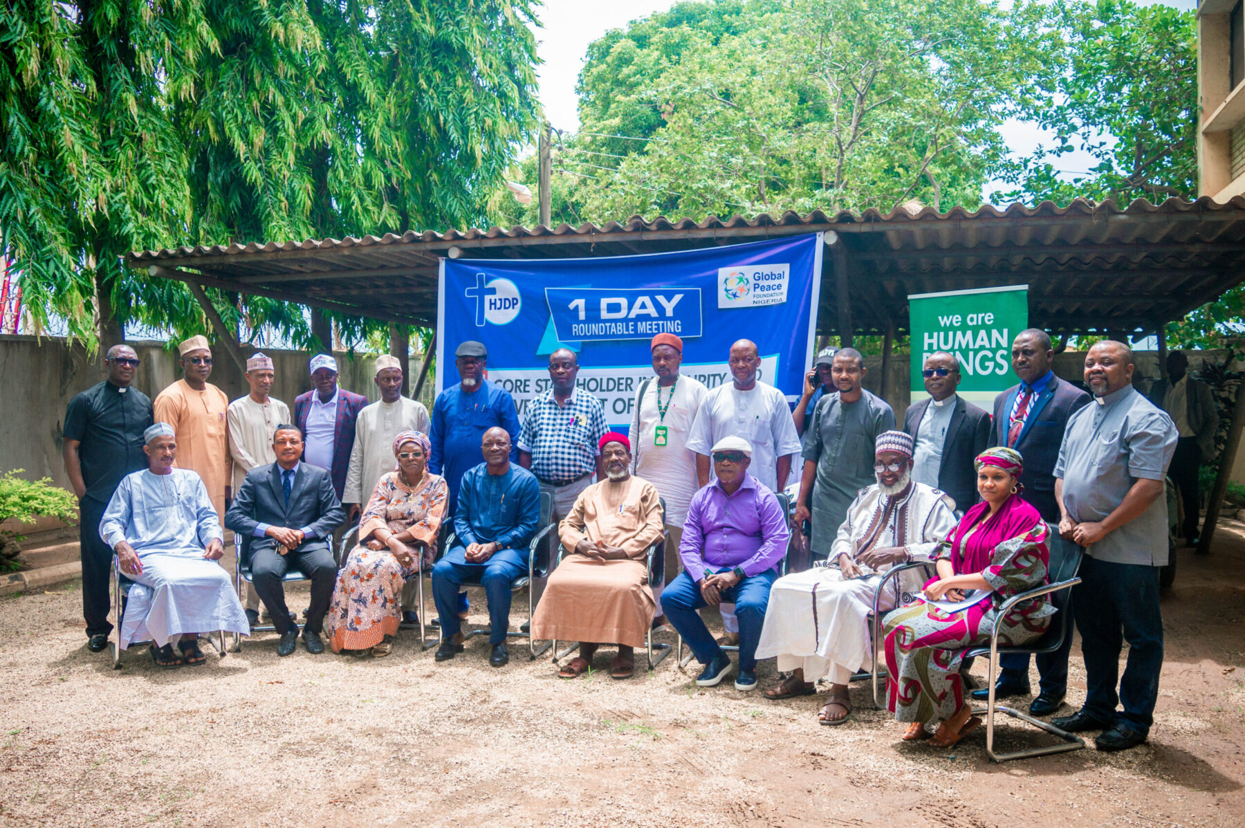 A group of diverse individuals posing for a photo in front of a banner during an outdoor stakeholder meeting, including government officials and religious leaders, with trees and a building in the background.