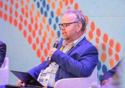 A person in a blue suit and glasses speaks into a microphone while holding a laptop. They are seated in front of a backdrop with an abstract orange dot pattern, discussing the Education Track for GPLC Africa 2024.