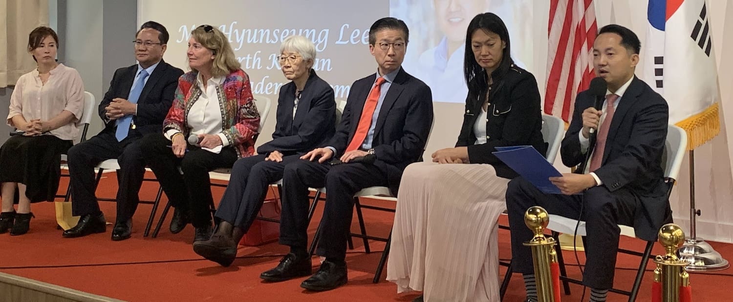 A group of seven people seated on a stage, some holding papers. There are American and South Korean flags in the background. A projected image and text are partially visible on the wall behind them, marking Defectors' Day at AKU Washington, highlighting North Korean defectors' stories.