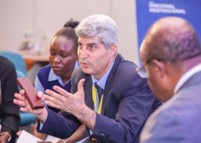 A man in a suit is speaking and gesturing with his hands in a group discussion. Two other people are engaged in the conversation, one taking notes, and another listening attentively, as part of the Education Track at GPLC Africa 2024.