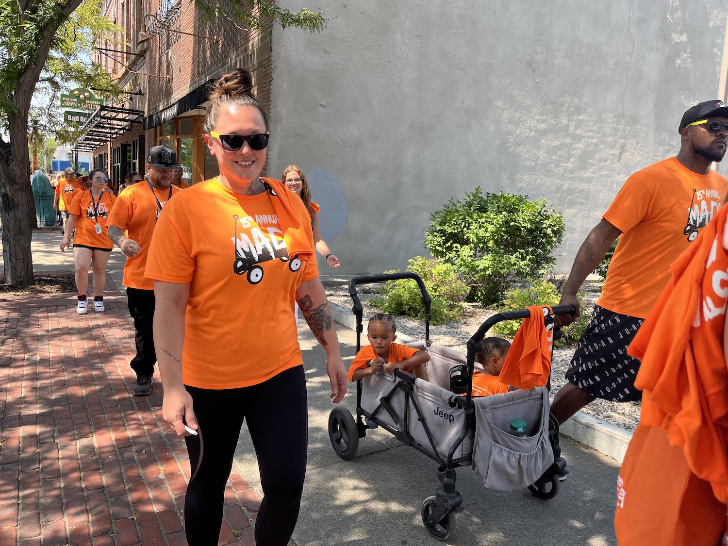 A group of people in orange T-shirts walk on a sidewalk during the Annual March. A woman pushes a double stroller with two children as they advocate against drugs and violence. Trees and a building are visible in the background, capturing a moment of unity within the USA community.