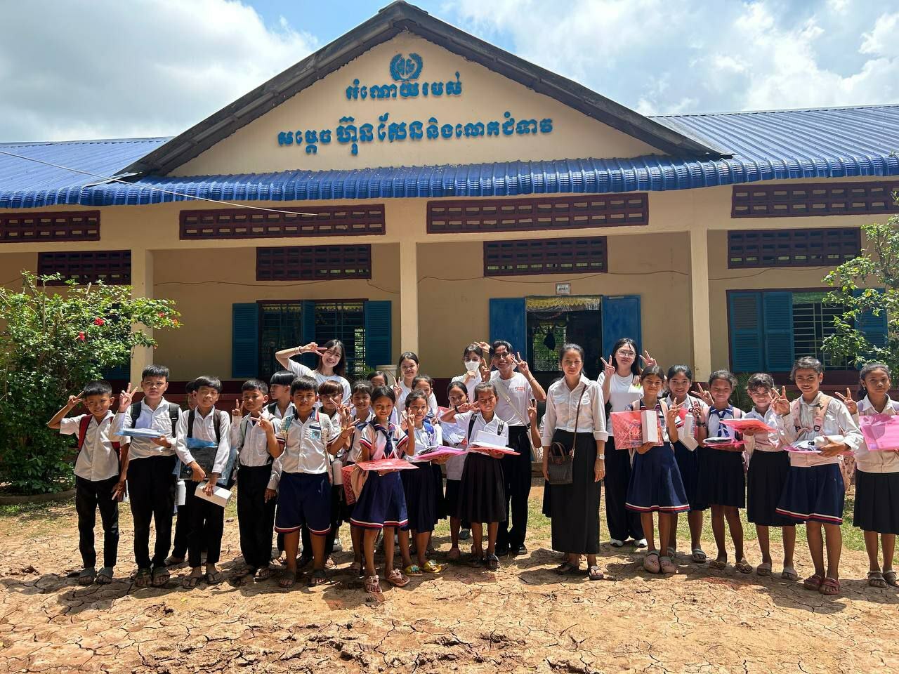 A group of students and teachers stand in front of a school building with blue roofing in Cambodia, holding gifts and supplies. Demonstrating youth leadership, they gather under the text written in the local language above the entrance.