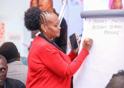 A person in a red sweater writes on a flip chart in a classroom or workshop setting. The chart reads "Primary partnership between schools & families." Others are present and attentive, clearly engaged in the Education Track at GPLC Africa 2024.