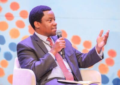 A man in a suit and striped tie speaks into a microphone during the Education Track at GPLC Africa 2024. He is seated on a white chair against a colorful background with orange and blue dots.