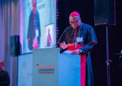 A clergy member speaks at a podium with "GPLC Africa 2024" displayed on the front during the plenary session. A large screen in the background shows his image. He reads from papers and gestures with one hand.
