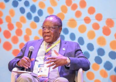 A man in a suit speaks into a microphone while seated at an event. A colorful polka-dot pattern adorns the background, and he wears a conference badge around his neck highlighting the Education Track at GPLC Africa 2024.