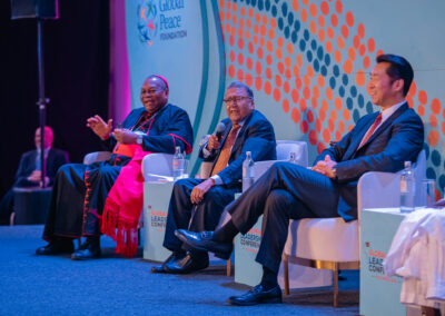 Three men sit on a stage with microphones and converse during the GPLC Africa 2024 Plenary. One wears a clerical robe, and the other two wear suits.