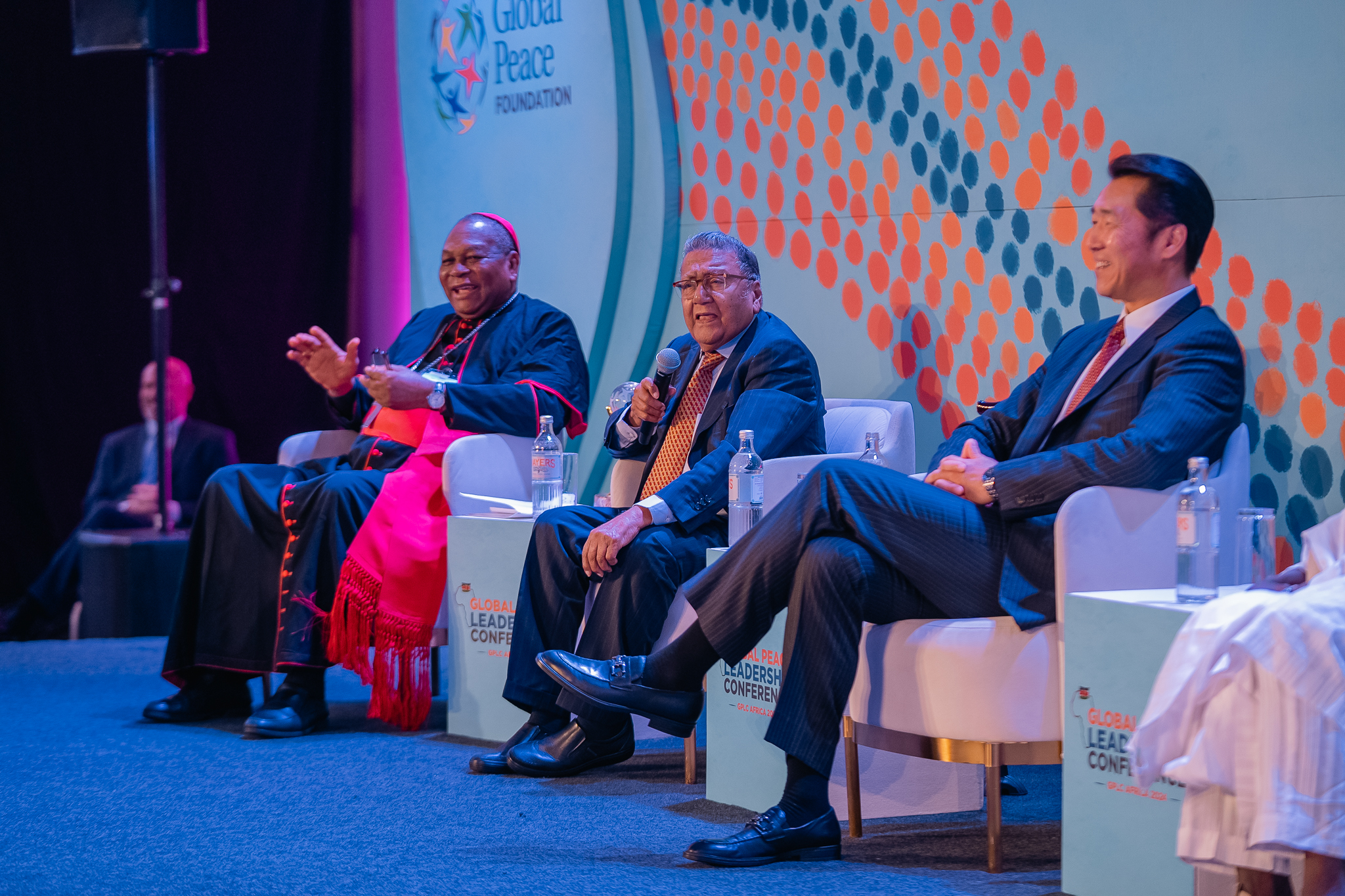 Three men sit on a stage with microphones and converse during the GPLC Africa 2024 Plenary. One wears a clerical robe, and the other two wear suits.