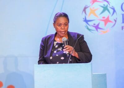 A person speaks into a microphone behind a podium during the GPLC Africa 2024 event. The colorful circle logo is displayed in the background on a blue wall, symbolizing the vibrant spirit of the Education Track.
