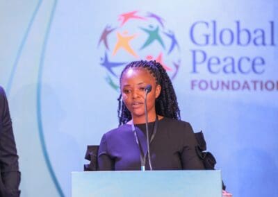A woman in a black dress speaks at a podium with microphones, with the "Global Peace Foundation" logo visible in the background. She passionately addresses the GPLC Africa 2024 Education Track, emphasizing the importance of transformative learning for future generations.