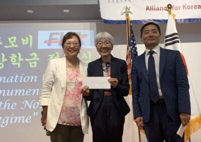Three people smile while a woman in the center holds an envelope. They stand in front of a screen with text, flanked by the American and South Korean flags, celebrating North Korean Defectors’ Day at AKU Washington.