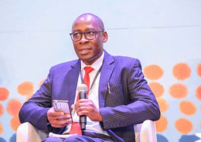 A man in a suit and tie speaks into a microphone while holding a smartphone, addressing the GPLC Africa 2024 Education Track, seated in front of a colorful backdrop.