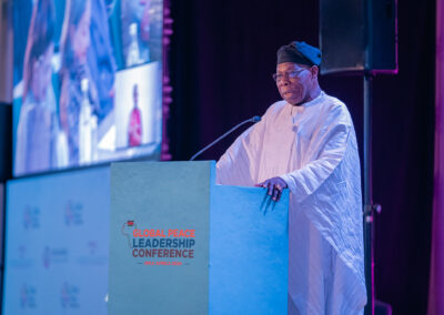 A person in traditional attire speaks at a podium labeled "GPLC Africa 2024." Multiple screens and a speaker setup are visible in the background, setting the stage for this significant plenary session at the Global Peace Leadership Conference.