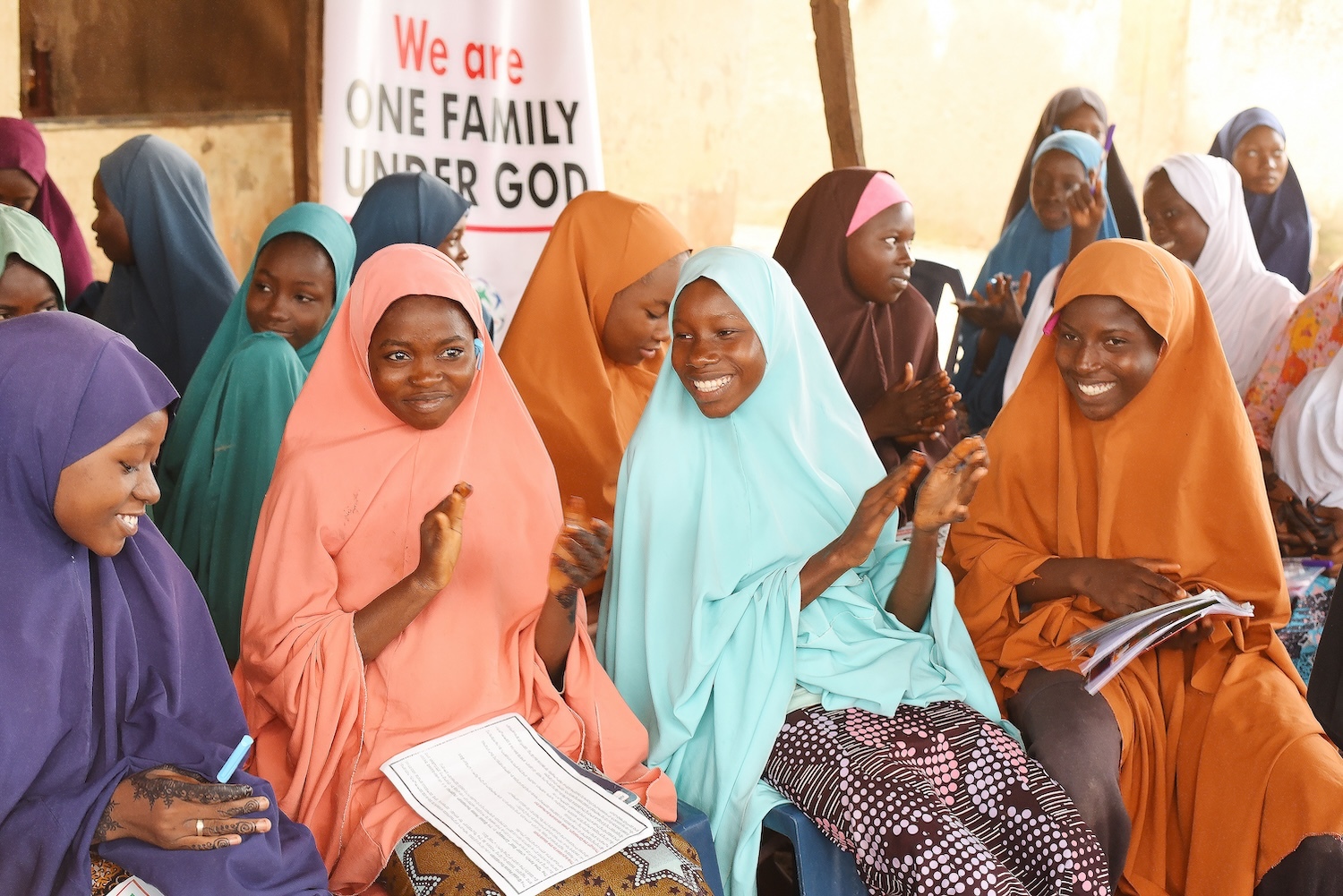A group of women in colorful hijabs sits together, some clapping and smiling, with a banner in the background reading "We are ONE FAMILY UNDER GOD," celebrating Healthy Living and Peace.