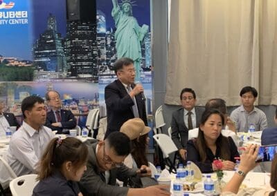 A man stands while speaking into a microphone in a room filled with several seated individuals, commemorating North Korean defectors. A backdrop shows the Statue of Liberty and cityscape, highlighting the significance of freedom. Tables with drinks and food are in the foreground, adding to the event's atmosphere.
