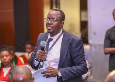 A man in business attire speaks into a microphone while standing in a crowd at GPLC Africa 2024. Several people are seated around him, listening attentively as he discusses the Education Track.
