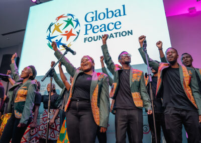 A group of people on stage wearing black clothing and patterned jackets raise their fists in the air in front of a "Global Peace Foundation" sign during the Plenary session at GPLC Africa 2024.