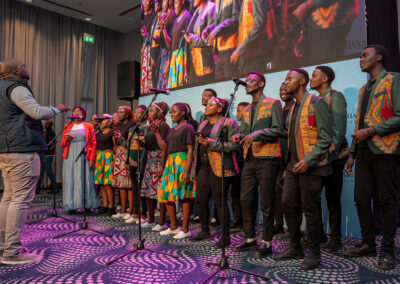 A choir dressed in vibrant clothing performs on stage with a conductor directing them, while a backdrop displays their image on a screen at the 2024 GPLC Africa Plenary.
