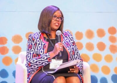A person with glasses, holding a microphone and papers, speaks while seated in front of a colorful dotted background, discussing the exciting Education Track planned for GPLC Africa 2024.
