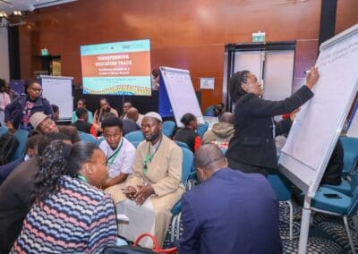 A diverse group of people engages in discussion around tables and whiteboards at the Transforming Education Track conference, part of GPLC Africa 2024. A presenter writes on a flip chart at the front of the room.