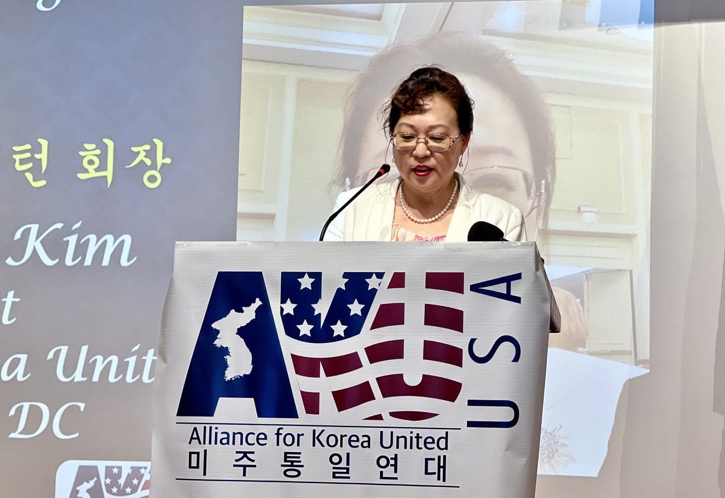 A woman stands at a podium speaking into a microphone. The podium features a banner reading