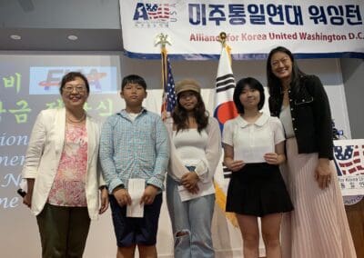 A group of four people stand in front of American and Korean flags, commemorating North Korean Defectors’ Day. A banner behind them reads "Alliance for Korea United Washington D.C." Two women are on the ends, with two children in the middle.