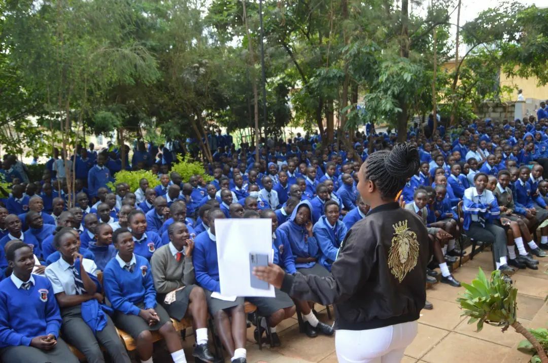 A woman is speaking to a large group of students wearing blue uniforms seated outdoors surrounded by trees. She holds a paper and a microphone, while the students listen attentively, eagerly awaiting to learn about the President’s Fellowship Program and various leadership opportunities available.