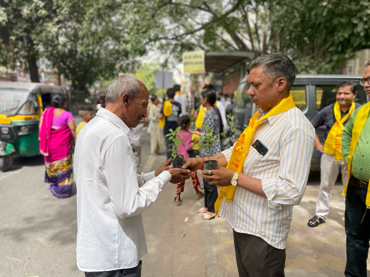 Two men exchange saplings on a busy street, celebrating World Environment Day. One man wears a yellow sash while people and auto-rickshaws bustle in the background. The event is part of an initiative by GPF India and JK Foundation to promote environmental awareness.