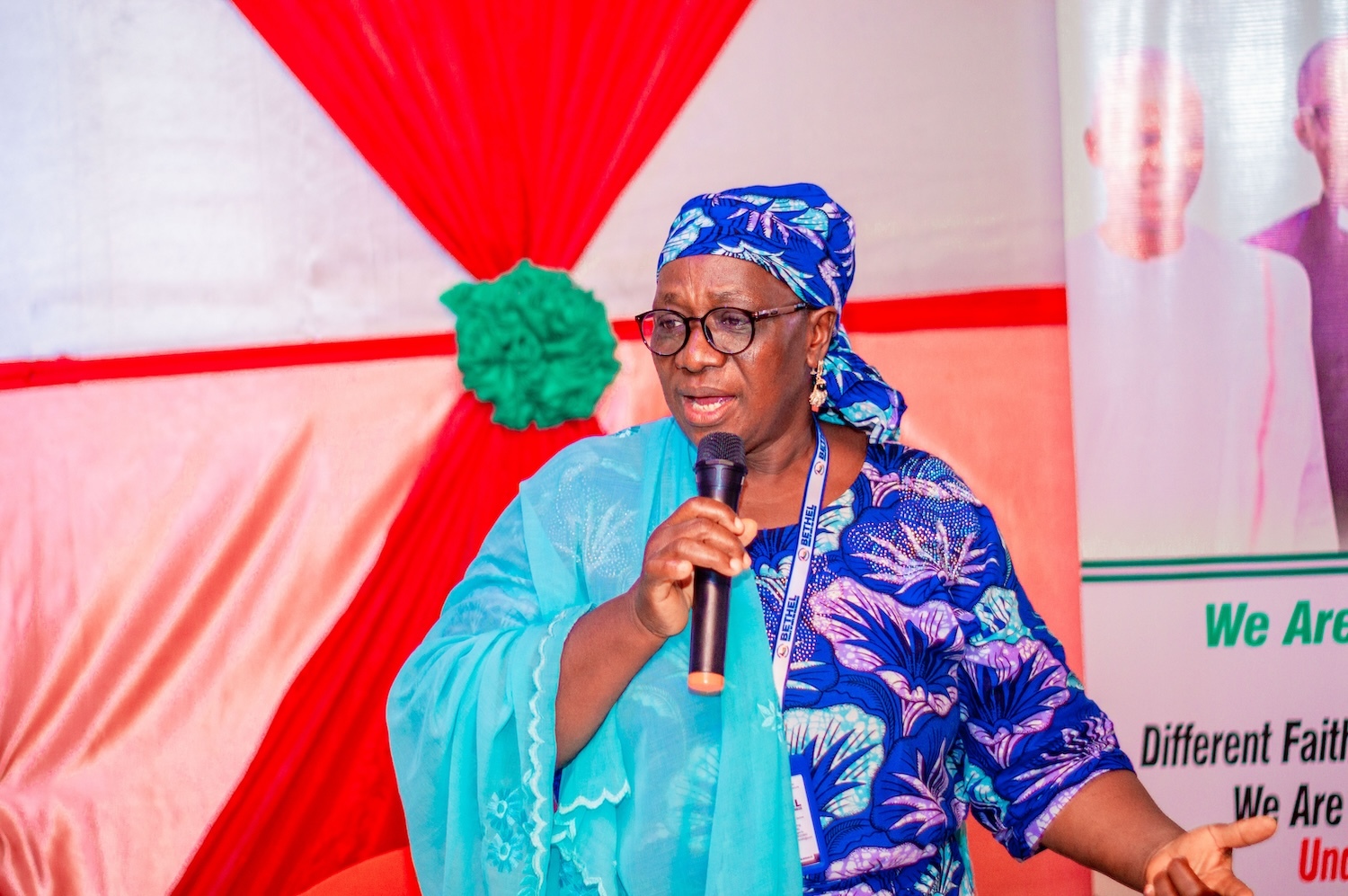 A woman in a blue outfit and headscarf speaks into a microphone, gesturing with her hand. She stands in front of a backdrop with red and white fabric and a Hayin Banki Kaduna banner, advocating for peace and reconciliation.