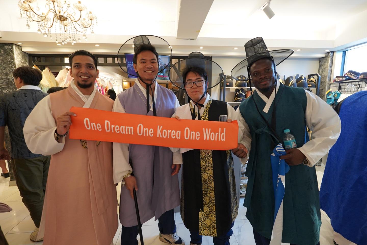 Four men wearing traditional Korean hanbok outfits hold an orange banner that reads "One Dream One Korea One World" in a brightly lit indoor setting.