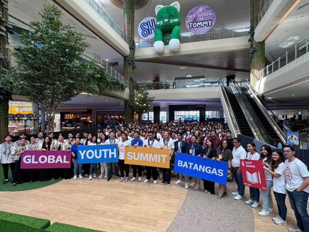 A large group of people gathered indoors, some holding signs that read “Global Youth Summit Batangas” and “Quality Education.” An escalator and a giant green plush toy are visible in the background. The event buzzes with discussions on building a sustainable future for the next generation.