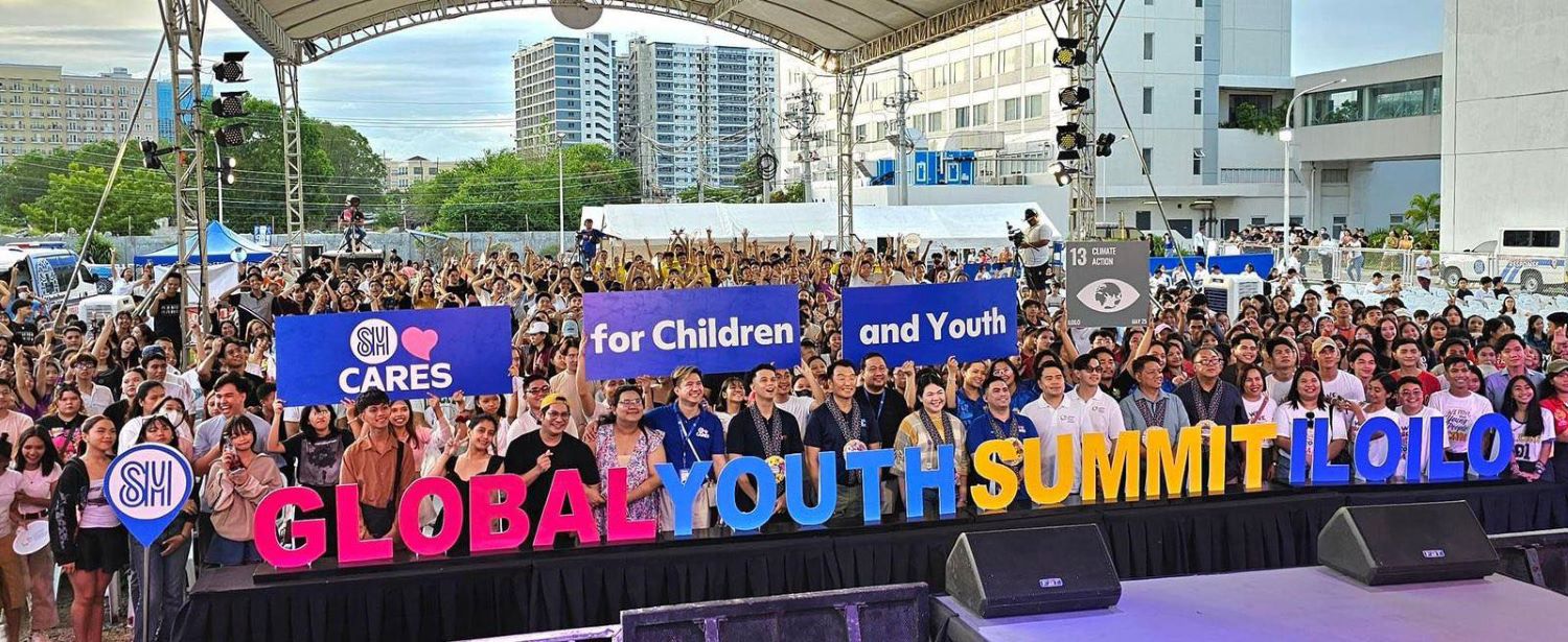 A large group of people stands on stage under a tent, holding signs that read "SM Cares," "for Children and Youth," and "Global Youth Summit Iloilo." An engaged audience is visible behind them, all committed to fostering a sustainable future for the next generation.