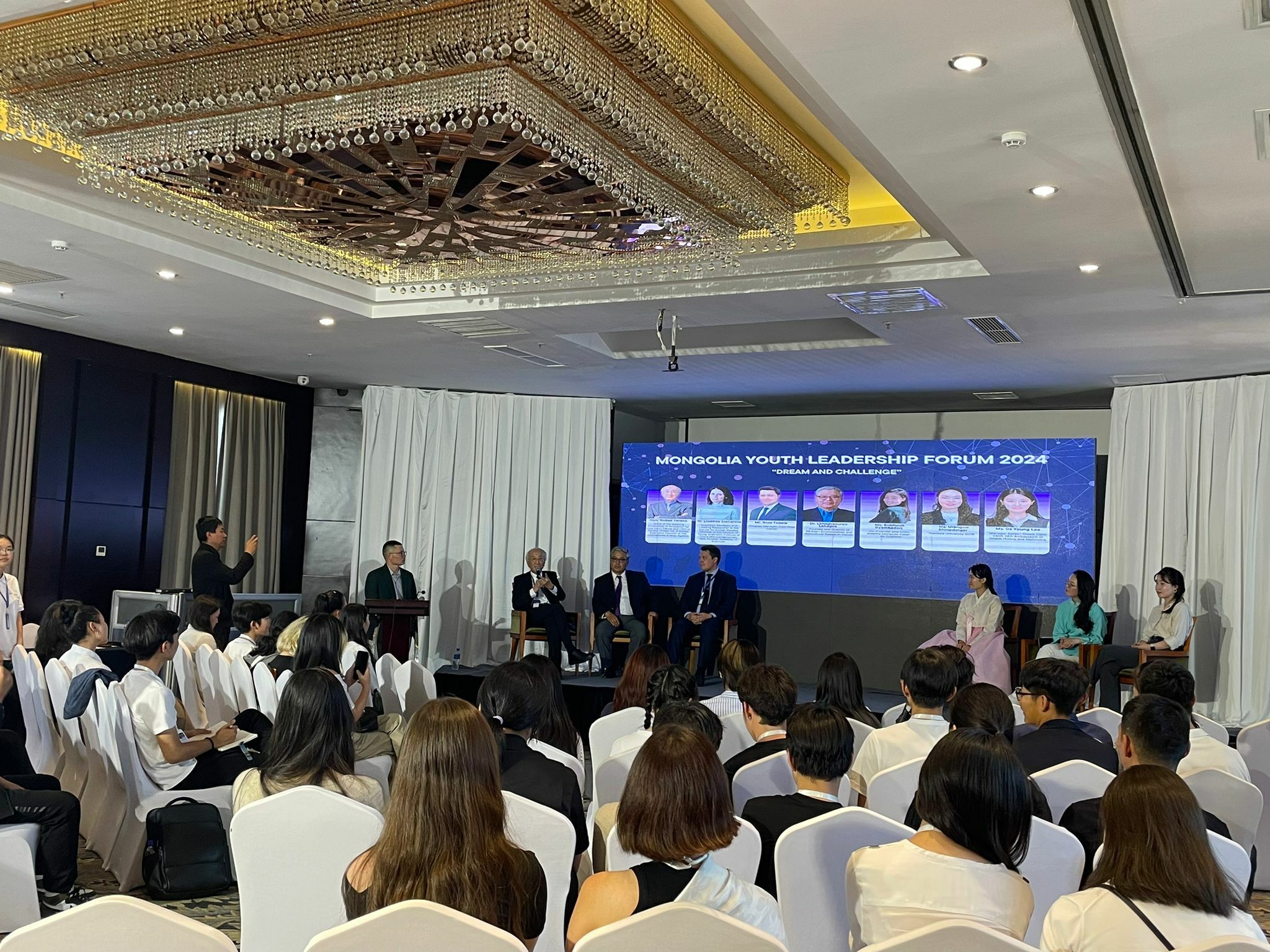 A group of people are sitting on a stage panel at the Mongolia Youth Leadership Forum 2023. An audience is seated facing them, and a large screen displaying information is behind the panelists. The event has garnered international attention, with Korean news outlets covering the discussions about regional collaboration.