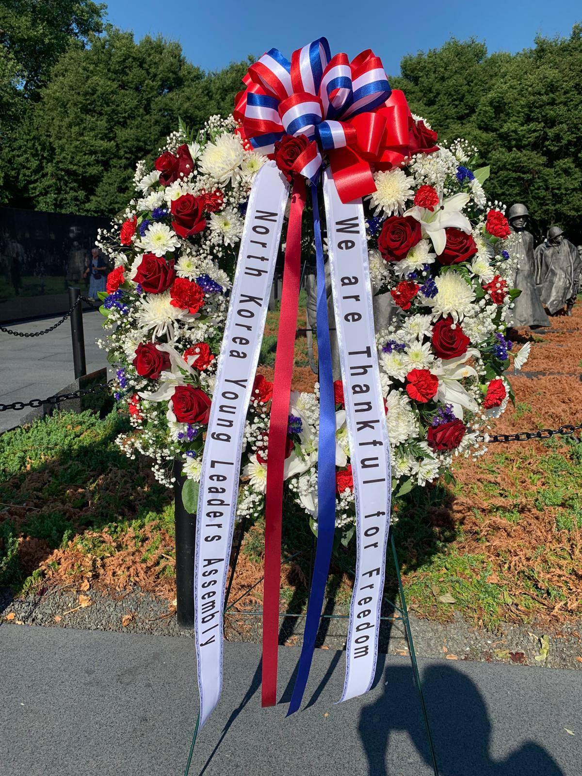 A floral wreath with red, white, and blue flowers and ribbons that read "North Korean Young Leaders Assembly" and "We are Thankful for Freedom" is displayed outdoors near the Korean War Memorial as a tribute.