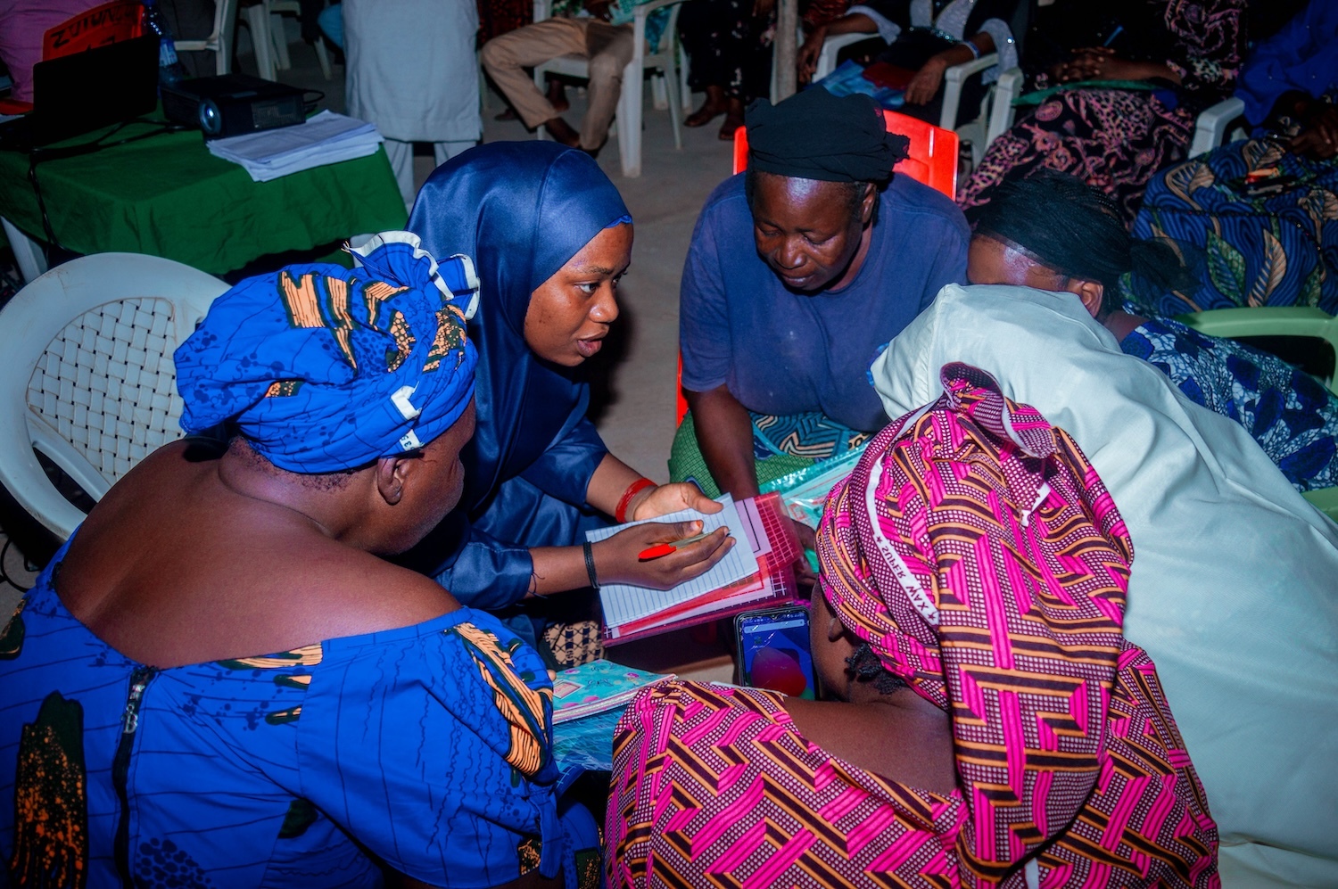 A group of women in colorful clothing sit together in discussion. One woman writes in a notebook while the others engage in conversation about interfaith peacebuilding efforts in Nigeria.