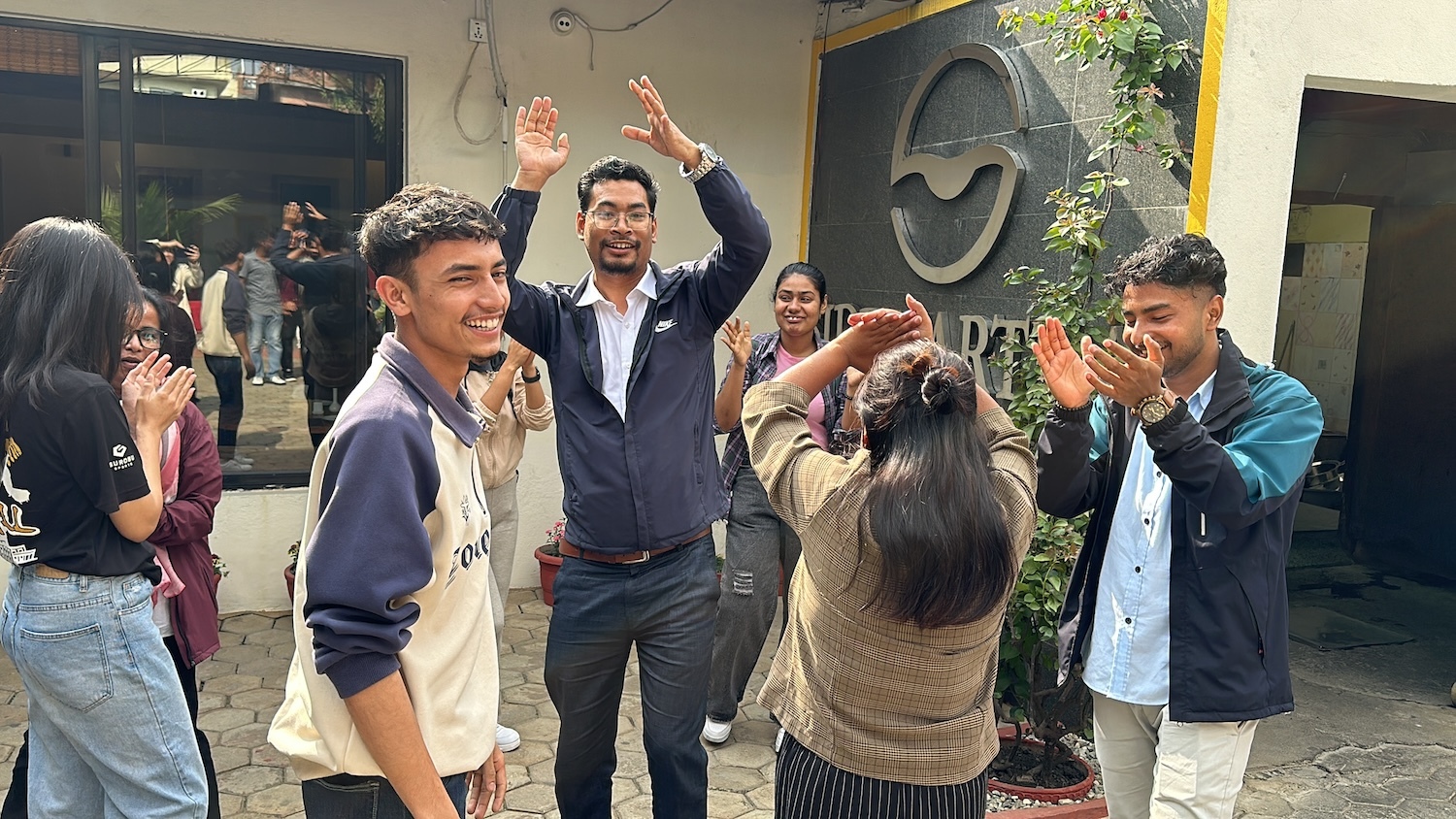 A group of people are standing outside, smiling and clapping. Some are raising their hands, and they appear to be celebrating or participating in a joyful activity together. Among them stand Nepali youth, embodying the spirit of innovative leaders shaping a brighter future.