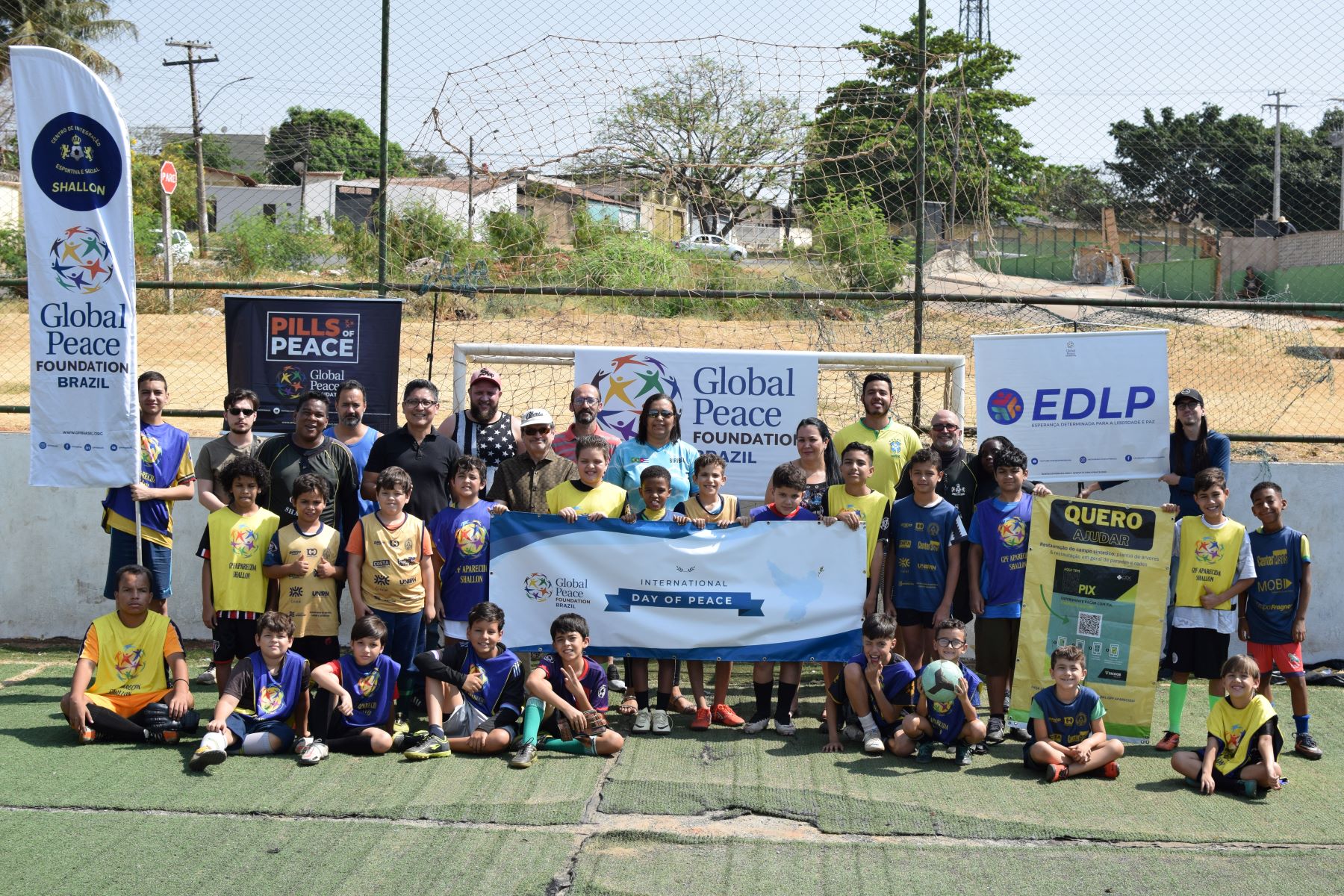 Group photo of children and adults on a soccer field in Aparecida de Goiânia, proudly holding an "International Day of Peace" banner. Youth in soccer attire kneel in front while the adults stand behind, with various promotional signs surrounding them.